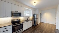This single-floor residence at Greybarn Amityville, located at 805 Broadway in Amityville, NY, offers a thoughtfully designed layout with modern finishes throughout. The kitchen is a standout feature, showcasing white shaker-style cabinetry with dark hardware, a classic white subway tile backsplash, dark countertops, and a full suite of stainless steel appliances including a range, microwave, dishwasher, and refrigerator. Under-cabinet lighting adds warmth, while a window above the sink brings in natural light. Rich, dark hardwood-style flooring flows through the kitchen and into the open living area, which is bathed in natural light from large windows overlooking the surrounding community. The spacious living area connects seamlessly to the rest of the unit, providing a bright and airy atmosphere. The bedroom features soft carpeting, generous wall space, and a window with neighborhood views, offering a comfortable and quiet retreat. The bathroom is finished with white subway tile surrounding a full soaking tub and shower, a white vanity with dark countertops, a large mirror, and classic mosaic tile flooring. In-unit laundry is also included. The overall design is clean, contemporary, and functional, making this a well-rounded living space.
