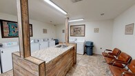 This shared laundry facility is designed to be both practical and welcoming, offering ample equipment and comfortable amenities in a single-floor layout. The space houses a substantial number of top-loading washers and dryers, each clearly numbered for easy identification and arranged along the perimeter walls to maximize efficiency and accessibility. Bright fluorescent lighting illuminates the entire room, ensuring a well-lit environment throughout the day.

A centrally positioned island serves as a focal point, featuring a granite-style countertop and a built-in utility sink, providing a convenient space for pre-treating garments or hand-washing delicate items. The island's rustic wood-paneled base adds a warm, decorative contrast to the otherwise neutral white walls and stone-patterned tile flooring that runs throughout the space.

Decorative wall art, including framed pieces with rustic and vintage aesthetics, adds character and visual interest to the room. A row of tan leather chairs lines one wall, offering a comfortable waiting area for residents. Trash receptacles are conveniently placed near the machines, and posted signage outlines facility rules and guidelines. A fire extinguisher and clearly marked exit ensure safety compliance throughout the space.