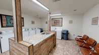 This shared laundry facility is designed to be both practical and welcoming, offering ample equipment and comfortable amenities in a single-floor layout. The space houses a substantial number of top-loading washers and dryers, each clearly numbered for easy identification and arranged along the perimeter walls to maximize efficiency and accessibility. Bright fluorescent lighting illuminates the entire room, ensuring a well-lit environment throughout the day.

A centrally positioned island serves as a focal point, featuring a granite-style countertop and a built-in utility sink, providing a convenient space for pre-treating garments or hand-washing delicate items. The island's rustic wood-paneled base adds a warm, decorative contrast to the otherwise neutral white walls and stone-patterned tile flooring that runs throughout the space.

Decorative wall art, including framed pieces with rustic and vintage aesthetics, adds character and visual interest to the room. A row of tan leather chairs lines one wall, offering a comfortable waiting area for residents. Trash receptacles are conveniently placed near the machines, and posted signage outlines facility rules and guidelines. A fire extinguisher and clearly marked exit ensure safety compliance throughout the space.
