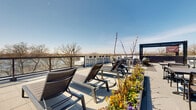 This expansive rooftop terrace is a thoughtfully designed outdoor amenity space offering sweeping, unobstructed views of a nearby waterway and surrounding tree line. The terrace is finished with large-format stone-effect pavers that lend a clean, modern aesthetic across the entire level. A glass and metal perimeter railing system preserves sightlines while ensuring safety, allowing residents and guests to take in the scenic waterfront panorama from virtually any point on the deck. The space is organized into several distinct zones, each designed for a different mode of outdoor enjoyment. A lounge area anchored by a bold navy steel pergola with strung bistro lighting offers a shaded retreat with plush sectional seating, ideal for relaxed gatherings. Adirondack chairs and a fire pit arrangement create a casual conversational setting, while rows of reclining sun loungers positioned along the railing invite sunbathing with direct water views. Bar-height seating along the perimeter provides additional options for alfresco dining or socializing. Raised planter beds filled with colorful seasonal blooms add warmth and natural beauty throughout the space. Abundant natural light floods the terrace throughout the day, and the open sky above creates a bright, airy atmosphere that makes this rooftop an exceptional outdoor destination.