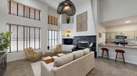 This thoughtfully designed single-floor space makes a striking first impression with its soaring double-height ceilings in the combined kitchen and living room. The living area is anchored by a floor-to-ceiling dark stacked-stone fireplace with a brass-trimmed insert, serving as a dramatic focal point. Built-in shelving flanks the fireplace and provides display space, while an oversized woven pendant light hangs above, adding an artisanal touch. Generous windows fitted with wood-slat blinds line the walls, flooding the space with natural light and enhancing the sense of openness created by the elevated ceilings. The upper wall features decorative art panels and a high shelf with greenery, adding visual interest at every level.

The kitchen is efficiently laid out with sage-green shaker cabinetry, stainless steel appliances including a range, microwave, and refrigerator, and stone-look countertops. A peninsula with bar seating provides a casual dining option and connects seamlessly to the living area. Warm hardwood-style flooring runs through the kitchen, complementing the carpeted living space.

The hallway offers additional room and connects the main areas, featuring high ceilings, natural light from well-placed windows, and space for additional furnishings. Throughout, the space maintains a cohesive, modern coastal aesthetic with neutral tones and natural textures.