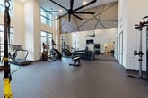 This fitness center is defined by its dramatic, soaring ceilings with exposed structural beams and large-scale ceiling fans that enhance air circulation throughout the space. Tall, multi-pane windows line the walls, flooding the interior with natural light and offering views of the surrounding exterior. The open floor plan is generous, providing ample room to move between equipment without feeling crowded.

The cardio zone features StairMaster steppers, ellipticals, and a treadmill positioned near the windows. The strength training area is thoroughly equipped with a full rack of dumbbells spanning a wide weight range, a Hoist functional training cable machine, a weight bench, medicine ball storage, and additional resistance machines. TRX suspension straps are anchored near the entrance column for bodyweight training versatility.

Full-length mirrors along one wall visually expand the space and aid in form monitoring. A wall-mounted flat-screen television is centrally positioned for easy viewing. The flooring consists of durable dark textured carpet tile throughout. A separate ancillary room is accessible through a doorway, and a water fountain is conveniently mounted near the entry. The overall ambiance is modern, clean, and purpose-built for serious fitness use.