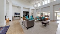 Located at 327 Guinevere Ln in Wilmington, NC, this single-story community clubhouse is designed to serve as a premier social hub. The space is anchored by a grand, open lounge area with soaring ceilings, large windows that invite abundant natural light, and light wood-style flooring throughout. Comfortable seating arrangements featuring gray sofas and teal accent chairs are arranged beneath a statement globe pendant light, creating an inviting atmosphere ideal for socializing and relaxing.

A dedicated game and lounge room offers a pool table, teal upholstered chairs, and direct access to an outdoor patio, bringing in views of the surrounding community. A separate media lounge provides dual wall-mounted televisions, a high-top bar counter with bar seating, and a striking textured wall mural as a focal point.

The full community kitchen is well-appointed with dark espresso cabinetry, stainless steel appliances, white quartz countertops, a large island with bar seating, and a window offering outdoor views. Adjacent to the main entrance, a coffee bar station with dark cabinetry and a commercial beverage machine provides a convenient amenity. The welcoming entry foyer features tall ceilings, large windows, and a stone accent wall with a mounted television, setting an impressive tone upon arrival.