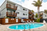 Located at 222 Columbia St in Santa Cruz, California, this outdoor amenity space serves as a central gathering hub within a multi-story residential community. The centerpiece of the courtyard is a generously sized, kidney-shaped swimming pool with crystal-clear blue water, complemented by a separate spa. The pool deck is finished with attractive stone-paver tile, providing ample space for lounging and socializing. White chaise lounge chairs and seating areas are arranged throughout the deck, along with a shaded dining area featuring a navy blue market umbrella and table-and-chair sets — ideal for outdoor relaxation.

The courtyard is enclosed by a combination of warm wood privacy fencing and black metal security fencing, creating a sense of seclusion while maintaining an open, airy feel. Mature palm trees rise throughout the space, lending a distinctly coastal California ambiance. Manicured hedges and tropical plantings line the perimeter, adding lush greenery to the setting.

Surrounding the pool area on multiple sides are the community's multi-story residential buildings, clad in grey siding and featuring wood-railed private balconies. Stone accent walls and exterior stairways contribute to the architectural character of the complex. The overall atmosphere is relaxed, resort-like, and well-suited to the sunny coastal climate of Santa Cruz.
