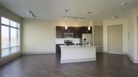 This single-floor space showcases a thoughtfully designed open-concept layout that seamlessly connects the kitchen and living areas. The kitchen is a standout feature, anchored by a large central island topped with granite countertops and equipped with an integrated sink. Dark espresso cabinetry lines the walls, providing generous storage while creating a striking contrast against the light-colored walls and white subway tile backsplash. Stainless steel appliances, including a French door refrigerator, gas range, and over-the-range microwave, lend a sleek, contemporary feel. Pendant lighting hangs above the island, complementing the track lighting that runs throughout the space, ensuring the area is well-lit from multiple angles. Rich, dark hardwood flooring extends across the entirety of the open floor plan, grounding the space with warmth and cohesion. The open living area offers generous room for furnishing and personalization, with neutral wall tones that provide a versatile backdrop. The overall ambiance is clean, modern, and functional, making this space well-suited for both everyday living and entertaining.