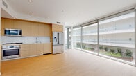 This thoughtfully designed one-bedroom, one-bathroom residence offers a clean, modern aesthetic across a single floor. The open-plan kitchen and living area is the heart of the home, anchored by expansive floor-to-ceiling glass windows and sliding doors that flood the space with natural light and frame views of a landscaped courtyard and surrounding architecture — with glimpses of palm trees visible from the bedroom.

The kitchen is outfitted with flat-front light wood cabinetry, a white quartz countertop with a marble-effect backsplash, and stainless steel Bosch appliances including a gas cooktop, built-in oven, microwave, and a french-door refrigerator. The living area extends generously from the kitchen, enhanced by light wood flooring that runs throughout.

The bedroom offers a calm retreat with direct access to the floor-to-ceiling glazing, a built-in closet with wire shelving, and light-toned hardwood floors. The bathroom is finished with warm-toned tile, a floating wood vanity with a white countertop, and a full soaking tub enclosed by a sliding frameless glass door. Recessed lighting and crisp white walls tie the space together, creating a bright and cohesive living environment.