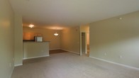 This single-floor space showcases an open-concept layout that seamlessly connects the living area to the kitchen. The main living area is finished with plush, neutral-toned carpeting, lending a comfortable and inviting atmosphere throughout the space. Warm beige walls complement the overall aesthetic, creating a cohesive and clean look.

The kitchen is positioned along one side and features a raised breakfast bar that serves as a natural divider between the cooking and living zones. Wooden cabinetry lines the upper portion of the kitchen, accompanied by a stainless steel appliance and a countertop with a darker finish. A pendant light fixture hangs above, providing focused illumination to the kitchen area, while a flush-mounted ceiling light brightens the living space.

A doorway visible toward the rear of the space leads to an additional room or closet area, suggesting further functional space beyond the main living zone. The flooring transitions from carpet in the living area to a harder surface near this back hallway. White baseboards and trim frame the rooms cleanly, adding a crisp finishing detail throughout the unit.
