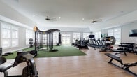 Located at 2700 Whakatane Ln in Fuquay-Varina, NC, this single-floor fitness center is a comprehensively outfitted facility designed to support a wide range of workout disciplines. The space is characterized by light-toned hardwood-style flooring, soft gray walls, recessed lighting, and ceiling fans that contribute to a bright and comfortable training environment. Abundant natural light filters in through multiple white-shuttered windows throughout, creating an open and airy atmosphere.

The main workout area is generously proportioned and divided into distinct functional zones. A turf-covered section serves as the hub for functional and strength training, housing a multi-station power rack with gymnastic rings, a punching bag, barbells, weight plates, and benches. Adjacent to this is a well-stocked cardio section featuring a row of treadmills, elliptical trainers, and stationary bikes by True Fitness, all positioned to face wall-mounted televisions.

A dedicated strength zone includes a cable machine, dumbbell rack, and adjustable bench, complemented by floor-to-ceiling mirrors that enhance both the visual spaciousness and training utility of the area. A separate studio room with mirrored walls, a ballet barre, spin bikes, and a full array of accessories including stability balls, medicine balls, resistance bands, and BOSU trainers supports group fitness and flexibility training. Additional leg press and selectorized machines round out the strength offerings, and a small retail or wellness nook is accessible from the main floor.