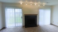 This two-story space features a well-appointed living area with several functional and aesthetic highlights. The main living space is finished with wall-to-wall carpet and centers around a striking fireplace clad in black tile, topped with a warm wood mantel that adds a natural contrast to the darker surround. Overhead, a track lighting system runs the length of the ceiling, providing adjustable, directed illumination throughout the room.

Natural light is a prominent feature of this space, with large windows dressed in vertical blinds flanking the fireplace. A sliding glass door on one side opens to a view of a grassy outdoor area, creating a strong visual connection to the exterior and allowing for ample daylight. The vertical blinds on both the door and the adjacent window offer flexible light and privacy control.

The walls are kept neutral, contributing to a clean and versatile backdrop suited to a variety of interior styles. The combination of the black tile fireplace, wood mantel, and track lighting gives the living area a balanced mix of modern and traditional elements. The space spans two floors, offering additional living or functional areas beyond what is visible.