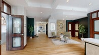 Located at 2551 38th Ave NE in St. Anthony, Minnesota, the Landings at Silver Lake Village community space is a generously sized, single-floor facility designed with both comfort and functionality in mind. Light-toned hardwood flooring flows consistently throughout, unifying the various areas and lending a warm, welcoming ambiance. The reception entry features a branded welcome mat, pendant lighting, and a front desk framed by wood-trimmed windows overlooking the exterior grounds. The expansive hallway connects the spaces seamlessly, with glass partition walls and sliding doors that maintain an open, transparent feel. Three distinct living areas each offer their own character: one showcases dramatic floor-to-ceiling arched windows draped in deep navy curtains with soaring ceilings and outdoor pool views; another features a natural stone accent wall, teal-painted accents, rich wood-framed interior doors, and sophisticated lounge seating; a third provides a more casual setting with flexible seating, a work counter, and cabinetry. A kitchen area includes a marble-topped island with bar seating, gray cabinetry, pendant lights, and a tile backsplash. Throughout, recessed lighting, bold accent walls, and curated greenery contribute to a polished yet inviting atmosphere.