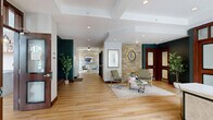 Located at 2551 38th Ave NE in St. Anthony, Minnesota, the Landings at Silver Lake Village community space is a generously sized, single-floor facility designed with both comfort and functionality in mind. Light-toned hardwood flooring flows consistently throughout, unifying the various areas and lending a warm, welcoming ambiance. The reception entry features a branded welcome mat, pendant lighting, and a front desk framed by wood-trimmed windows overlooking the exterior grounds. The expansive hallway connects the spaces seamlessly, with glass partition walls and sliding doors that maintain an open, transparent feel. Three distinct living areas each offer their own character: one showcases dramatic floor-to-ceiling arched windows draped in deep navy curtains with soaring ceilings and outdoor pool views; another features a natural stone accent wall, teal-painted accents, rich wood-framed interior doors, and sophisticated lounge seating; a third provides a more casual setting with flexible seating, a work counter, and cabinetry. A kitchen area includes a marble-topped island with bar seating, gray cabinetry, pendant lights, and a tile backsplash. Throughout, recessed lighting, bold accent walls, and curated greenery contribute to a polished yet inviting atmosphere.