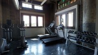 Located at 340 SW 5th St in Des Moines, Iowa, this fitness facility offers a well-equipped workout space designed for both functionality and comfort. The room features a dark, durable flooring surface suited for high-traffic exercise use, complemented by exposed brick walls and dark-trimmed windows that lend an industrial character to the space. Multiple large windows line the walls, flooding the room with natural light and providing open views of the surrounding cityscape — creating an energizing atmosphere for workouts. The ceiling height adds a sense of openness, and the overall layout is thoughtfully arranged to maximize usable floor space. Cardio equipment, including treadmills and an elliptical trainer, is positioned to take advantage of the natural light and views. A dumbbell rack with free weights is also available, supporting a range of strength training needs. The combination of raw architectural elements — including visible brick and industrial ceiling details — with modern fitness equipment gives the space a distinctive, urban aesthetic. Natural light, city views, and a spacious layout make this a practical and motivating environment for fitness activity in the heart of Des Moines.