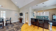 This single-floor clubhouse in Cary, NC, is designed as a sophisticated communal gathering space with a thoughtfully curated layout and upscale finishes throughout. The centerpiece of the main lounge is a floor-to-ceiling stacked stone fireplace with a wall-mounted television above, framed by bold navy blue accent walls and patterned drapery panels that filter natural light from multiple windows. Vaulted ceilings soar overhead, amplifying the sense of openness and volume throughout the space. Comfortable teal and blue striped sofas and accent chairs are arranged around a dark wood coffee table, creating an inviting seating area ideal for relaxed gatherings. Adjacent to the lounge, a wet bar area features dark granite countertops, glass-front cabinetry, a decorative mosaic tile backsplash, and pendant lighting. High-top bar seating provides a casual dining option, while nearby bistro tables with leather chairs offer additional seating near large windows overlooking greenery outside. The flooring transitions seamlessly between warm hardwood and richly patterned carpet tiles, defining distinct functional zones. A workstation area is also visible within the space, adding practical utility. Overall, the clubhouse balances a refined aesthetic with a welcoming, community-focused atmosphere.