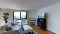 This single-floor space showcases a thoughtfully designed living area with a clean, contemporary aesthetic. The living room is anchored by a sleek, wall-mounted linear fireplace that serves as a stylish focal point, complemented by a large flat-screen television atop a dark wood media dresser. White upholstered seating, including a sofa, armchair, and matching ottoman, are arranged atop a patterned blue area rug, creating a cohesive and inviting seating arrangement. Warm hardwood flooring extends throughout the space, adding natural texture and warmth to the otherwise neutral palette. Large floor-to-ceiling windows with black frames allow generous natural light to fill the room, offering views of the surrounding neighborhood and glimpses of palm trees, suggesting a California or similarly temperate US location. Recessed ceiling lighting supplements the natural light, ensuring the space remains bright throughout the day. The white walls and minimalist design approach contribute to an open, airy atmosphere that feels both modern and comfortable. Overall, the space presents a well-balanced blend of functional furnishings and stylish architectural details.