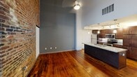 This two-story apartment at 1101 Hawthorne Lane in Charlotte, NC blends industrial character with modern finishes. The main floor features rich hardwood flooring, dramatic soaring ceilings, and a striking exposed brick accent wall. The open kitchen includes granite countertops, dark cabinetry, and stainless steel appliances with pendant lighting. A staircase leads to the upper level, where both bedrooms offer hardwood floors and spacious walk-in closets. One upper bedroom showcases an exposed wood-plank ceiling with a window for natural light. Three bathrooms include granite vanities and a combination tub-shower. The overall ambiance is a stylish urban loft aesthetic.