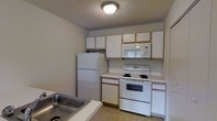 This single-floor space features a functional kitchen designed with efficiency in mind. The kitchen is equipped with a full suite of white appliances, including a top-freezer refrigerator, an electric coil range with oven, an over-the-range microwave, and a dishwasher integrated beneath the counter. White cabinetry with warm wood-tone trim lines the upper walls, providing ample storage space above the countertops. The countertops are light-colored and extend to accommodate a stainless steel sink, positioned conveniently near the workspace. The layout follows a practical U-shaped or galley-style configuration, making efficient use of the available space. Light tile flooring anchors the room, complementing the neutral gray walls and white cabinetry. A flush-mount ceiling light fixture provides general illumination throughout the kitchen. The textured ceiling adds a subtle design element consistent with the overall understated aesthetic of the space. The kitchen's clean, straightforward design prioritizes functionality, offering all the essential elements for everyday cooking and meal preparation within a well-organized, compact footprint.