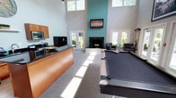 This impressive single-floor amenity lounge is designed for both comfort and entertainment, showcasing a dramatic double-height ceiling that amplifies the sense of openness throughout the space. The room's most striking architectural feature is a floor-to-ceiling teal accent wall housing a classic fireplace and a wall-mounted flat-screen TV, serving as a bold focal point that draws the eye upward toward clerestory windows that bathe the interior in abundant natural light.

The layout is thoughtfully divided into distinct activity zones. A full-sized billiards table anchors the central area, providing a dedicated recreation space, while a cozy lounge area with plush armchairs and a side table is positioned near the fireplace, creating an inviting spot for relaxation and conversation. Textured, patterned wallcoverings in soft gray tones add depth and sophistication to the walls.

A kitchenette along one side of the room features dark countertops, wood cabinetry, and appliances, offering convenient amenities for gatherings and events. Multiple sets of glass French doors line the perimeter, connecting the interior to outdoor areas and allowing natural light to pour in from multiple directions. Ceiling fans positioned throughout ensure year-round comfort, while large framed artwork contributes to the space's polished, community-oriented atmosphere.