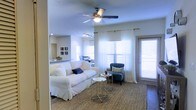 This single-floor residence offers an inviting and well-lit living space designed for comfortable everyday living. The main living area is anchored by a plush white slipcovered sofa paired with a navy blue accent chair, creating a classic yet casual seating arrangement. A woven area rug grounds the space, while a pair of sleek metallic side tables add a modern touch. Sheer white curtains frame multiple windows, including a glass door, allowing generous natural light to flood the interior and create an airy, open atmosphere. A ceiling fan with an integrated light fixture is centrally positioned overhead, providing both function and airflow. Track lighting in the background adds supplemental illumination throughout. The open layout flows naturally into a dining area visible in the background, where a dining table and chairs are set beneath additional windows. A partial view of the kitchen reveals granite or stone countertops and cabinetry beyond. The dark hardwood-style flooring adds warmth and contrast to the light-toned walls, tying the overall aesthetic together cohesively.