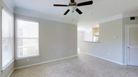 This single-story space features an open-concept layout that connects the living area to the kitchen, creating a cohesive and functional flow throughout. The living area is finished with plush wall-to-wall carpeting in a neutral tone, complemented by crisp white baseboards and crown molding that add a polished architectural detail along the ceiling perimeter. A ceiling fan with a built-in light fixture is centrally mounted, providing both air circulation and ambient lighting. A window along the side wall allows natural light to filter into the space, brightening the room's warm, neutral color palette.

The kitchen is visible through a pass-through counter opening, which enhances the open feel of the layout while maintaining a defined cooking area. The kitchen features light-toned cabinetry, a refrigerator, and overhead lighting that keeps the space bright and functional. The pass-through counter also offers potential for use as a breakfast bar or casual dining area. Overall, the space presents a clean, understated aesthetic with consistent neutral finishes throughout, making it adaptable to a variety of interior styles.