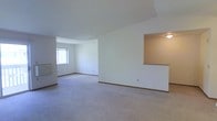 This single-floor space presents an open, airy layout characterized by clean white walls and neutral-toned wall-to-wall carpeting throughout. The main living area is generously proportioned, with a vaulted or angled ceiling detail that adds architectural interest and enhances the sense of vertical space. A window along one wall admits natural light, brightening the interior and contributing to the overall open feel.

A defining feature of this space is the pass-through opening cut into one of the interior walls, which connects adjacent areas and promotes an open flow between rooms. This architectural detail, framed with a warm wood-toned ledge, also allows light to travel between spaces. A ceiling-mounted light fixture is visible through the opening, providing supplemental illumination to the adjoining area.

The layout includes a secondary room or alcove visible through a wide entryway, offering flexibility for use as a bedroom, office, or additional living space. Warm wood baseboard trim runs consistently along the base of the walls, providing a subtle contrast to the light flooring and white walls. Overall, the space conveys a simple, functional aesthetic with a clean, move-in-ready presentation.