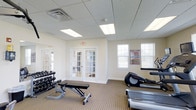 This fitness center is a dedicated, single-floor facility designed to support a comprehensive workout routine. The space is finished with a neutral-toned carpet and warm beige walls, creating a clean and welcoming environment for exercise. A drop ceiling with recessed fluorescent panel lighting ensures the room is consistently and evenly lit, while multiple windows fitted with horizontal blinds allow natural light to filter in, enhancing the overall brightness of the space.

The facility is equipped with a range of exercise equipment, including a treadmill, an elliptical machine, and a rowing machine for cardiovascular training. A rack of dumbbells in varying weights, paired with an adjustable weight bench, provides options for strength and resistance training. A workout reference chart is mounted on the wall, offering a convenient visual aid for exercise guidance.

Access to the fitness center is provided through a set of elegant French doors with glass panels, which open to an adjacent area. An emergency exit sign is clearly visible, reflecting attention to safety standards. The overall layout is open and functional, making efficient use of the available floor space while maintaining an uncluttered, easy-to-navigate environment suitable for a variety of fitness activities.