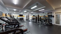 This fitness center is a comprehensive, single-floor facility designed to support a full range of workout routines. The space is defined by a bold visual identity, featuring a striking yellow accent stripe running along the walls and vivid athletic murals — depicting dynamic silhouettes of runners, weightlifters, and athletes — that energize the environment and reinforce its purpose. The flooring throughout consists of durable dark rubber matting, ideal for both cardio and strength training activities. A row of Star Trac treadmills lines one wall, each equipped with individual display screens for entertainment during workouts. Adjacent to these, a bank of elliptical trainers provides additional low-impact cardio options. The strength training area is outfitted with a variety of selectorized machines and cable systems, supported by large floor-to-ceiling mirrors that enhance the sense of space and allow for form checks. A dumbbell rack and flat benches provide versatility for free weight training, while a rowing machine adds another functional fitness option. The ceiling features a coffered design with recessed and linear fluorescent lighting that ensures the space is brightly and evenly illuminated. Windows along one wall allow natural light to filter in, complementing the overall clean and motivating atmosphere of this well-appointed fitness facility.
