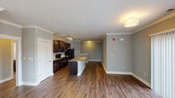 This two-floor residence is finished with consistent neutral gray walls and warm hardwood-style flooring throughout the main living areas, creating a cohesive and polished aesthetic. The open-concept main floor centers around a well-equipped kitchen outfitted with rich dark wood cabinetry, granite countertops with a tile backsplash, black appliances including a range, microwave, and refrigerator, and a central island with a sink. Track lighting illuminates the kitchen, which flows seamlessly into the living area. The living room is generously proportioned and features crown molding, a ceiling fan, and a sliding glass door flanked by additional windows that invite abundant natural light and outdoor views. Bedrooms are carpeted for comfort, with multiple windows providing natural light and ventilation. The bathrooms are well-appointed, one featuring a glass-enclosed tiled shower, and another with a dark wood vanity topped with granite and a window overlooking greenery. A dedicated laundry room accommodates a full-size washer and dryer side by side, with a window and built-in shelving above for added convenience.