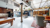 Located at 700 Everett St in Richmond, VA, this single-floor space offers an expansive, open-plan layout with a thoughtfully curated industrial-modern aesthetic. The architecture retains its raw character through exposed structural timber posts, white-painted wood beam ceilings, and overhead ductwork, all of which contribute to an airy, loft-like atmosphere with impressive ceiling height throughout.

The floor plan is organized into distinct functional zones while maintaining a seamless, flowing feel. A lounge area features mid-century-inspired wingback chairs, a cognac leather sofa, a glass coffee table, and a plush area rug, complemented by a wall-mounted shelving unit and a large built-in wooden bookcase. Pendant globe lighting and a brass chandelier add warmth overhead. A communal dining and social zone offers high-top bar tables with industrial stools alongside smaller café-style tables, ideal for group gatherings. A kitchen area with dark cabinetry and full-size appliances supports the space's event and coworking capabilities. A pool table provides recreational interest, while a private phone booth pod offers a quiet retreat. Deep slate-blue accent walls contrast effectively with white ceilings and light concrete flooring, and lush hanging plants throughout bring vibrant organic energy to the space.