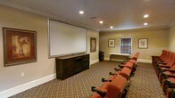This single-floor screening room is thoughtfully designed to deliver a true cinematic experience. At the heart of the space is a large wall-mounted projection screen, paired with a dark wood media console beneath it that houses audiovisual equipment. A row of plush, rust-colored theater-style seats with built-in armrests and cup holders lines the room, arranged to provide excellent sightlines to the screen. The patterned carpet in warm earth tones adds a sense of comfort and absorbs sound, enhancing the room's acoustic quality. Warm, neutral-toned walls complement the classic interior, which is accented by crown molding and recessed lighting that allows for flexible illumination control. Framed artwork adorns the walls, adding a refined touch to the cinematic setting. A window with slatted blinds provides the option for natural light while maintaining blackout capability for optimal viewing. A side table along the back wall offers additional functional space. The overall ambiance is inviting and sophisticated, blending the comfort of a living space with the functionality of a professional screening room.