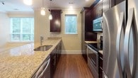 This single-floor, 2-bedroom, 2-bathroom residence offers a thoughtfully designed layout with consistent, high-quality finishes throughout. The kitchen is a standout feature, showcasing rich dark espresso cabinetry, granite countertops, and a full suite of stainless steel appliances including a refrigerator, range, and built-in microwave. A curved island with a undermount sink and pendant lighting above serves as both a functional workspace and a natural transition into the open living area. The living space is generously proportioned, with large windows on multiple walls that invite abundant natural light and offer views of surrounding greenery. Crown molding and hardwood flooring add a refined touch to the main living and kitchen areas. Both bedrooms are carpeted and well-lit, with the primary bedroom benefiting from corner window placement for added brightness and a walk-in closet. Each bathroom mirrors the home's design aesthetic with dark vanity cabinetry, granite countertops, and frameless glass shower enclosures. One bathroom features a shower-tub combo, while the other offers a dedicated walk-in shower. Warm neutral tones carry throughout the entire residence, creating a cohesive and welcoming atmosphere.