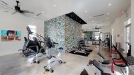 This well-appointed fitness center is designed with both functionality and aesthetics in mind. The space is anchored by a dramatic freestanding mosaic-clad structure at its center, finished in a multicolored tile pattern blending teal, brown, and neutral tones. A dark ceiling panel caps the structure, creating a bold architectural focal point that draws the eye throughout the room.

The flooring throughout is polished concrete, providing a durable and clean surface well-suited to a high-traffic fitness environment. Recessed lighting is installed across the bright white ceiling, ensuring consistent, even illumination throughout the space. A ceiling fan is visible toward the rear, contributing to airflow and comfort during workouts.

The equipment layout is thoughtfully organized. Cardio machines, including stationary bikes and a StairMaster, are positioned toward the front of the space. Free weights, benches, and strength training equipment are arranged along the sides and toward the rear, with large mirrors lining the walls to support proper form. Natural light filters in through windows along the perimeter, enhancing the open, energetic atmosphere of this clean and contemporary fitness facility.
