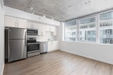 This single-floor urban unit showcases a compelling blend of industrial aesthetics and modern design. Exposed concrete ceilings run throughout the space, lending a raw, loft-like character that is balanced by crisp white walls and light-toned wood-style flooring underfoot.

The open-concept kitchen is well-appointed with a full suite of stainless steel appliances, including a refrigerator, electric range, over-the-range microwave, and dishwasher. White cabinetry with brushed metal hardware provides ample storage, while track lighting mounted to the concrete ceiling adds both function and visual warmth.

A sliding barn door connects the living area to an interior room, adding a stylish architectural detail while maximizing usable space. The bedroom benefits from large windows that draw in natural light and offer views of the surrounding urban streetscape, creating a bright and airy atmosphere.

The bathroom is cleanly designed with light wood-toned cabinetry, a single-sink vanity with a white countertop, a large mirror, and elegant pendant lighting. Wood-look tile flooring ties the space together seamlessly.

The 3D floorplan reveals a thoughtfully laid-out unit with a logical flow between the living, kitchen, bedroom, and bathroom areas. Overall, the space combines the character of industrial architecture with the comfort and practicality of contemporary finishes.