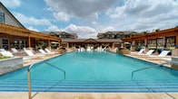This expansive resort-style pool area is designed to deliver a premium outdoor living experience. The centerpiece is a large, sparkling swimming pool with vivid turquoise water, framed by a broad travertine pool deck lined with white lounge chairs for sunbathing. Distinctive modern in-water chaise loungers invite residents to relax directly in the shallow pool shelf, adding a cabana-style touch to the amenity.

Surrounding the pool, a series of wooden pergolas provide shaded gathering spaces fitted with plush wicker furniture accented by vibrant blue cushions, drapery panels, and additional patio seating beneath large market umbrellas. A stone fire pit with Adirondack chairs creates a cozy focal point for cooler evenings.

The covered outdoor kitchen area is equipped with multiple stainless steel built-in grills set into a limestone-clad counter, ideal for outdoor entertaining. Nearby, large shade canopies shelter hammock areas and casual seating overlooking a well-maintained green lawn. Lush landscaped planters with colorful seasonal plantings accent the pool perimeter, adding a vibrant natural element. A limestone tower serves as a notable architectural landmark anchoring the community clubhouse, which frames the backdrop of the pool area alongside multi-story residential buildings.