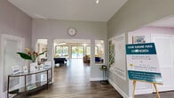 Located at 4120 Union Square Blvd in Palm Beach Gardens, Florida, this single-floor community clubhouse offers a generous and thoughtfully designed gathering space. The layout flows seamlessly across multiple interconnected living areas, all finished with durable wood-look flooring and a neutral, contemporary color palette throughout.

The welcoming entry foyer features a console table, community information displays, and open archways that frame views straight through to the pool area beyond, flooding the space with natural light. The main lounge area boasts tray ceilings with recessed lighting, large sliding glass doors opening to the pool deck, and comfortable seating arrangements that invite relaxation.

A second lounge area includes a pool table, foosball table, and additional seating, creating a lively entertainment zone. Adjacent to this is a kitchenette with pendant lighting, a central island with bar stools, and stainless steel appliances. A cozy reading nook features a bookshelf accent wall and plush armchair, offering a quiet retreat. The multi-purpose community room provides a long communal table, bar-height seating along the windows, and vibrant artwork, ideal for gatherings or remote work. Soft pink accent walls near the entry corridor add a cheerful touch to this well-appointed community hub.