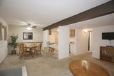 This single-floor home offers a well-organized layout with a variety of functional living spaces. The living room serves as the heart of the home, furnished with two plush, dark upholstered sofas accented with decorative pillows, neutral carpet flooring, and a window with draped blinds that allows natural light to filter in. A decorative beam detail adds architectural character to the ceiling. Adjacent to the living area, the dining space accommodates a round wooden table with matching chairs and ceiling fan overhead, with a pass-through window connecting to nearby rooms and adding a sense of openness. The compact kitchen is efficiently designed, equipped with a coil-burner range and a full-size refrigerator alongside overhead cabinetry. The bedrooms are carpeted and each feature ceiling fans with light fixtures. The primary bedroom includes dual closets and a wooden dresser, while a secondary bedroom is versatile enough to serve as a children's room or flex space, complete with a window for natural light. A full bathroom features a tub-shower combination with a bold striped curtain, a vanity with storage, and tile flooring. A lower-level media room with exposed beam detail, wall-mounted television, and access to additional storage rounds out this practical and comfortable home.