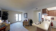 This single-story space showcases an open-concept layout that combines the living, dining, and kitchen areas into one cohesive, functional environment. The living area is anchored by plush wall-to-wall carpeting that adds warmth and comfort underfoot, while neutral-toned walls create a clean, versatile backdrop throughout.

A standout feature is the set of French doors that flood the living area with abundant natural light, offering direct access to the outdoors and enhancing the sense of openness. A framed piece of wall art adds a decorative touch near the doors, complementing the relaxed atmosphere.

The kitchen transitions seamlessly into the living space via a breakfast bar-style counter, finished with a light countertop surface that doubles as a casual dining area. Dark wood cabinetry visible above adds contrast and a touch of warmth to the kitchen zone. A secondary doorway, framed in clean white trim, connects to an additional room, suggesting a well-organized and accessible floor plan. Overhead, ceiling vents indicate a central HVAC system for year-round comfort. Overall, the space delivers a bright, practical, and inviting atmosphere.