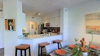This well-appointed two-floor residence showcases a thoughtfully designed interior with modern finishes and a clean, contemporary aesthetic. The open-concept kitchen serves as a central highlight, featuring crisp white cabinetry that extends to the ceiling, providing generous storage. A mosaic tile backsplash adds a decorative focal point, complementing the granite countertops that wrap around the breakfast bar. The bar seating area, lined with upholstered stools, creates a natural transition to the dining space, making the layout ideal for both everyday living and entertaining. Stainless steel appliances, including a refrigerator and built-in microwave, complete the kitchen with a polished, functional feel. Track lighting illuminates the space efficiently, enhancing the bright, airy atmosphere created by the light-toned walls and cabinetry. The dining area adjacent to the kitchen offers a dedicated space for meals, furnished with a full dining table and coordinating chairs. The layout flows seamlessly across two floors, offering distinct zones for living and private quarters. Overall, the space presents a cohesive, modern design with attention to detail, practical functionality, and a welcoming ambiance suited for contemporary living.