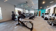 This expansive fitness center is thoughtfully designed to support a wide range of workout styles across distinct training zones. The cardio area is outfitted with a comprehensive lineup of equipment, including treadmills, ellipticals, stationary bikes, and stair climbers, several featuring interactive digital displays. Large glass-panel garage doors open fully to integrate the interior with an outdoor turf area, where additional equipment such as benches, weight racks, and a heavy bag provide space for functional and outdoor training. A dedicated strength and resistance zone houses a variety of selectorized machines arranged along mirrored walls and large windows, creating an open, well-lit environment. A separate functional training room features a rubber floor, a wall-mounted rig with suspension trainers and resistance bands, a tire, and a BOSU ball, catering to high-intensity and cross-training workouts. Distinctive blue vertical slat dividers define zones while maintaining visual openness throughout the space. Vibrant teal accent walls, dark ceiling treatments with recessed and linear lighting, and patterned carpet flooring contribute to a modern, energetic aesthetic. Ceiling fans and ample natural light from multiple window configurations further enhance the overall training environment.