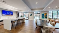 This expansive single-floor amenity lounge offers a thoughtfully designed open-concept layout that seamlessly blends multiple functional zones into one cohesive, inviting space. Rich hardwood-style flooring flows throughout, unifying each area beneath crisp white ceilings with recessed lighting and decorative troffers that provide warm, even illumination.

The main lounge area anchors the space with a sleek tile-surround linear fireplace, a large wall-mounted TV, and comfortable seating arranged over a plush area rug — ideal for relaxed gatherings. Adjacent to this, a game area features a billiard table and shuffleboard, while oversized windows with black metal framing flood the room with natural light and provide views of an outdoor terrace.

The wet bar and kitchenette are fully equipped with taupe shaker-style cabinetry, white quartz countertops, a stainless steel sink, under-counter refrigerator, and a wall-mounted TV above — making it well-suited for catering and entertaining. A long bar island with pendant lighting offers additional seating via sleek counter-height stools.

A semi-private dining nook features marble-topped bistro tables surrounded by upholstered seating and striking wall art panels, creating an intimate breakout setting. Throughout, curated artwork in vivid blues and warm tones adds a contemporary artistic character to the overall space.