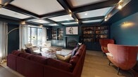 This thoughtfully designed single-floor space makes a bold architectural statement through its rich, deep teal and near-black color palette carried consistently throughout. The living area serves as the centerpiece, featuring a striking coffered ceiling with integrated track lighting and exposed dark beams that add depth and dimension overhead. Built-in shelving units flank a marble-surround fireplace, while a generously sized burgundy sectional sofa and rust-colored accent chairs create an inviting, layered seating arrangement. Wide-plank hardwood flooring runs throughout, anchoring the space with warmth.

The gourmet kitchen is exceptionally well-appointed, showcasing teal-painted cabinetry with brass hardware, a dramatic stone slab backsplash, a professional-grade gas range, and a panel-ready refrigerator. A substantial butcher block island with an undermount sink and bar seating serves as both a functional workspace and a social hub. Pendant lights with brass fixtures hang above, complementing the warm wood tones.

The formal dining area offers an intimate yet refined atmosphere, centered around a large rectangular table with upholstered chairs for generous seating. A tiered gold chandelier commands attention overhead, while textured wall treatments and sheer drapery-lined windows contribute to the room's polished, gallery-like ambiance.