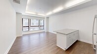 Located at 10 Dorrance St in the heart of Providence, Rhode Island, this single-floor unit offers three bedrooms, two bathrooms, and a thoughtfully designed layout that maximizes its compact footprint. The open-concept living and kitchen area serves as the central hub of the space, featuring warm hardwood-style flooring, recessed lighting, and a distinctive tray ceiling with integrated ambient lighting that adds architectural depth. Large windows in the living area allow natural light to filter in, providing views of the surrounding urban streetscape.

The kitchen is outfitted with white shaker-style cabinetry, granite countertops, stainless steel appliances including a refrigerator and range, and a central island that offers additional prep and storage space. Two hallways connect the living areas to the three bedrooms, each finished with neutral gray walls, hardwood-style flooring, and clean white trim. One bedroom includes a built-in closet, enhancing storage functionality.

Both bathrooms are finished to a high standard, featuring white subway tile walling, penny-tile flooring, granite-topped white vanities, and brushed gold fixtures that add a refined touch. The overall aesthetic throughout the unit is clean, modern, and cohesive, with a neutral palette that creates a bright and functional living environment.