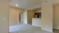 This single-floor unit features an open-concept layout that seamlessly connects the living area to the kitchen. The living space is finished with wall-to-wall carpeting in a light neutral tone, contributing to a clean and understated aesthetic. Walls are painted in a warm beige, creating a cohesive and inviting atmosphere throughout. Recessed lighting in the ceiling provides functional illumination, complemented by a ceiling-mounted light fixture. A hallway visible from the main living area leads to additional rooms, with two white panel doors offering access to separate spaces, suggesting a bedroom and closet or bathroom nearby. The kitchen is defined by a raised breakfast bar with a light-toned countertop, providing a natural boundary between the cooking and living zones. Dark espresso-toned upper cabinets add contrast against the neutral walls, while a stainless steel refrigerator anchors the kitchen's appliance lineup. Tile or hard flooring transitions into the kitchen area, distinguishing it from the carpeted living space. Ceiling-mounted HVAC vents are present throughout, ensuring efficient climate control. Overall, the space presents a functional and well-organized interior with a straightforward, modern residential design.
