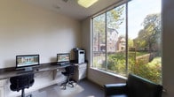 This business center is a single-floor workspace thoughtfully designed to support productivity and convenience. The room features a built-in granite-topped counter that runs along the walls, providing ample workspace for multiple users simultaneously. Two computer workstations are set up side by side, each equipped with a monitor and keyboard, alongside a multifunction printer and copier for comprehensive office needs. Overhead cabinetry with a clean, white finish offers generous storage, keeping the space organized and functional. The flooring is covered in a neutral-toned carpet, and recessed and flush-mounted ceiling lights ensure consistent, even illumination throughout. A standout feature of this space is the large floor-to-ceiling corner window that floods the room with natural light while offering picturesque views of lush greenery, flowering plants, and surrounding buildings. A comfortable black leather armchair is positioned near the window, providing a relaxed seating option. A glass-paneled interior door connects to an adjacent corridor, enhancing accessibility. The overall ambiance is clean, professional, and welcoming, making it a practical and comfortable environment for work-related tasks.