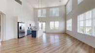 Located at 921 N Central Ave in Tampa, FL, this contemporary two-story residence showcases an impressive open-concept design anchored by soaring double-height ceilings that flood the living area with natural light through two tiers of large windows. Light wood-style flooring extends across the main living and kitchen spaces, enhancing the bright, airy ambiance. The kitchen is a standout feature, outfitted with a two-tone cabinet scheme pairing crisp white upper cabinets and rich navy-blue lower cabinets, all accented with brushed gold hardware. A clean white quartz countertop, glossy subway tile backsplash, stainless steel appliances including a French door refrigerator, range, and microwave, along with a central island, complete this well-appointed space. The living area benefits from sweeping outdoor views of the Tampa skyline and surrounding greenery. Bedrooms are carpeted for comfort, with ceiling fans and ample natural light. One bedroom offers direct views of the city skyline. The primary suite includes a glass-enclosed walk-in shower with white tile surround and an adjacent walk-in closet. Bathrooms feature wood-toned hardware, white cabinetry, and light wood flooring, maintaining a cohesive, modern aesthetic throughout.