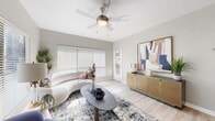 This thoughtfully designed single-floor residence combines modern aesthetics with functional living across several well-defined spaces. The living room serves as the heart of the home, featuring light hardwood-style flooring, neutral gray walls, and large windows dressed with white horizontal blinds that invite an abundance of natural light. A ceiling fan adds both comfort and character, while a curved white sofa, an organic-shaped dark coffee table, and a gold media credenza topped with large-scale abstract artwork create a sophisticated, curated atmosphere. A dedicated dining area offers an intimate setting with a round marble-top pedestal table, vibrant orange accent chairs, and colorful wall art — a cheerful and functional space for meals and gatherings. The bedroom is a serene retreat, highlighted by sliding glass doors opening to a private balcony with outdoor views, a ceiling fan, and light flooring. A dramatic dark upholstered headboard with chevron detailing anchors the room, complemented by bold orange bedding and a walk-in closet. The bathroom features large-format stone-look tile surrounding a soaking tub and shower combination, a clean white vanity, and understated contemporary décor, completing this cohesive and well-appointed residence.