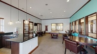 This single-floor community clubhouse is thoughtfully designed to offer a variety of recreational and social amenities in one open, well-appointed space. The main gathering area is generously sized, featuring high ceilings with elegant crown molding, recessed lighting, and a combination of tile and patterned carpet flooring that defines different activity zones.

At the heart of the space is a standout kitchenette anchored by a large island with a sleek dark granite countertop, dark wood cabinetry, and a sink — illuminated by a trio of globe pendant lights that add a modern touch. Multiple windows with wood-slatted blinds allow ample natural light to fill the room.

A full-size billiards table serves as a centerpiece of the game area, complemented by casual seating arrangements including round tables and leather chairs. A vibrant teal accent wall defines a cozy lounge nook equipped with a wall-mounted television and intimate bistro-style seating — ideal for games or casual conversation.

Classic dark wood French doors with glass pane inserts connect to a private room furnished with a desk, leather chair, wall-mounted TV, and a cue rack, offering a quieter retreat. The dollhouse view also reveals a dedicated fitness area with exercise equipment positioned near large windows, completing this versatile and inviting communal facility.