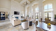 Located at 7145 Reading Rd in Rosenberg, TX, this single-floor community clubhouse offers a generously sized, open layout designed to impress. The grand living area commands attention with dramatically soaring ceilings, elegant coffered detailing, and stately arched doorways that lend a sense of architectural refinement. A floor-to-ceiling stacked stone fireplace serves as the focal centerpiece, topped by a mounted flat-screen television, and flanked by neutral-toned sofas and accent chairs arranged for comfortable gathering. Natural stone tile flooring runs throughout, complementing the light, airy color palette. A contemporary chandelier adds a sophisticated touch overhead. Expansive floor-to-ceiling windows and glass entry doors flood the space with abundant natural light while framing views of the outdoors. A dedicated dining area with dark wood tables provides additional seating. The galley-style kitchen is outfitted with dark granite countertops, a bold mosaic tile backsplash, stainless steel appliances, and a breakfast bar with bar seating — a stylish and functional space for entertaining. A billiards table is also visible, adding a recreational element. Wall sconces and recessed lighting ensure the space is well-lit throughout, creating a welcoming, upscale ambiance.