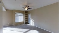 This single-floor space features a well-lit main room finished with wall-to-wall carpeting and neutral-toned walls that create a warm, understated ambiance. A ceiling fan with an integrated light fixture is centrally mounted, providing both air circulation and overhead lighting. Natural light streams generously through multiple windows fitted with horizontal blinds, casting bright patterns across the floor and enhancing the open feel of the room. A set of built-in shelves lines one corner, offering practical and permanent storage or display space without occupying additional floor area. A paneled door with a slatted window insert leads to an adjoining space, adding architectural interest while allowing light to filter through. The clean lines, soft carpeting, and functional built-ins contribute to a livable and versatile interior. The overall layout appears straightforward and efficient, suited to a variety of uses. The combination of natural light, built-in storage, and ceiling fan makes this a practical and comfortable single-level space.