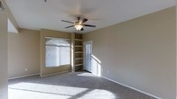 This single-floor space features a well-lit main room finished with wall-to-wall carpeting and neutral-toned walls that create a warm, understated ambiance. A ceiling fan with an integrated light fixture is centrally mounted, providing both air circulation and overhead lighting. Natural light streams generously through multiple windows fitted with horizontal blinds, casting bright patterns across the floor and enhancing the open feel of the room. A set of built-in shelves lines one corner, offering practical and permanent storage or display space without occupying additional floor area. A paneled door with a slatted window insert leads to an adjoining space, adding architectural interest while allowing light to filter through. The clean lines, soft carpeting, and functional built-ins contribute to a livable and versatile interior. The overall layout appears straightforward and efficient, suited to a variety of uses. The combination of natural light, built-in storage, and ceiling fan makes this a practical and comfortable single-level space.
