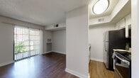 This single-floor residence offers a thoughtfully laid-out living space with a clean, neutral aesthetic throughout. The living area serves as the heart of the home, featuring rich dark hardwood-style flooring and a sliding glass door fitted with vertical blinds that floods the room with natural light while providing access to an outdoor view of lush greenery. A dedicated entry door and built-in wall panel add functional character to this main space.

The galley-style kitchen is efficiently designed, offering white cabinetry, granite-style countertops, and a full suite of stainless steel appliances including a refrigerator, range, microwave, and dishwasher. Light wood-style flooring ties the kitchen seamlessly to the adjacent hallway and common areas.

A central hallway connects the main living zone to the private spaces, including a bedroom finished with plush carpet flooring, a ceiling fan with light fixture, and a generously sized window that brings in abundant natural light and outdoor views. The bathroom features a white vanity with a granite-topped countertop, framed mirror, and bright overhead lighting, with additional storage visible beyond.

Throughout the residence, textured ceilings, crisp white walls, and consistent wood-style flooring create a cohesive and comfortable atmosphere well-suited for everyday living.